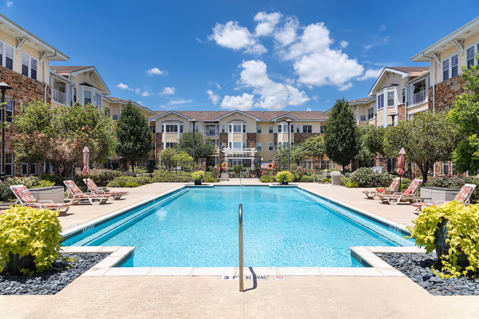Outdoor swimming pool with lounge chairs flanked by multi-story apartment buildings and landscaping under a blue sky.