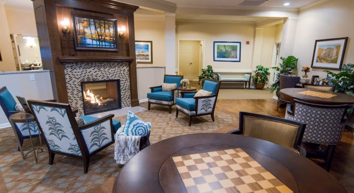 A cozy senior living facility common area with a lit fireplace surrounded by four upholstered chairs. There are two round tables with chess/checkerboard patterns and chairs around them. The room is decorated with framed artwork, plants, and soft lighting, creating a warm and inviting atmosphere.