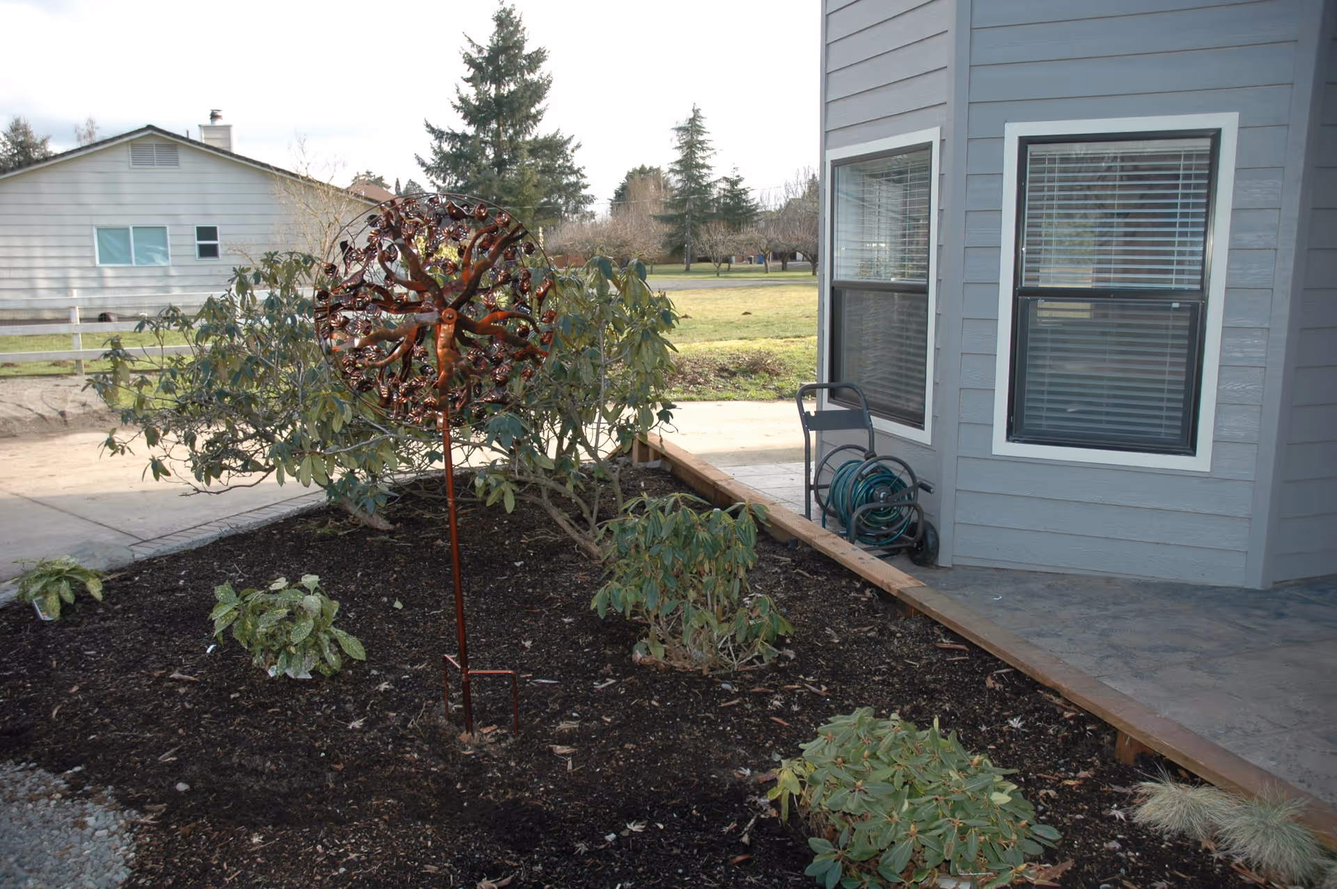 Outdoor garden area next to a building with gray siding and two windows. The garden has several small plants and a decorative metal sculpture shaped like a tree. A green garden hose is coiled on a cart near the building. In the background, there is a lawn, trees, and another house with light-colored siding.
