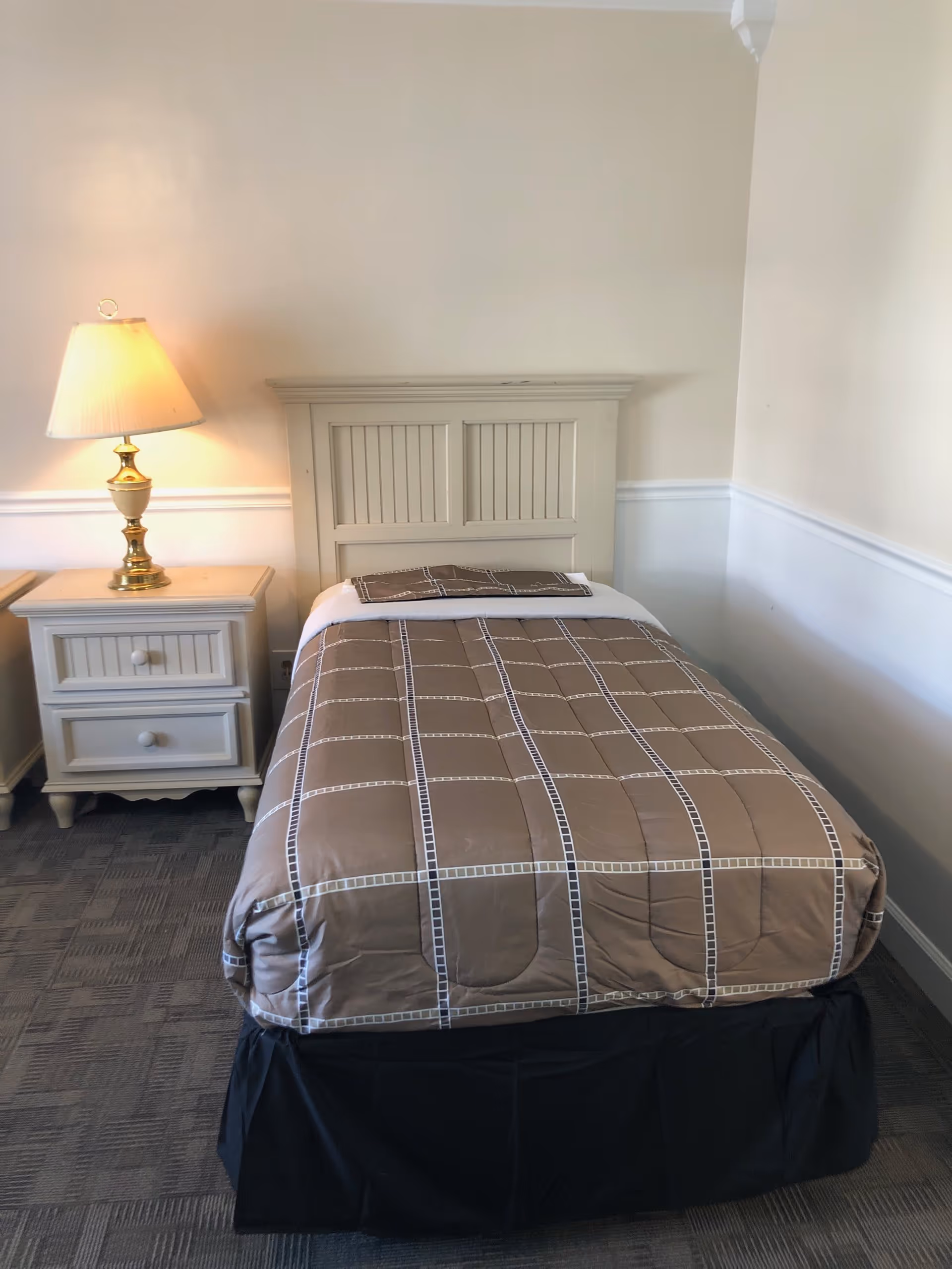 A single bed with a beige and brown checkered comforter and a matching pillow is placed against a light-colored wall. Next to the bed is a white nightstand with two drawers and a brass lamp with a beige lampshade. The floor is carpeted with a patterned gray carpet.