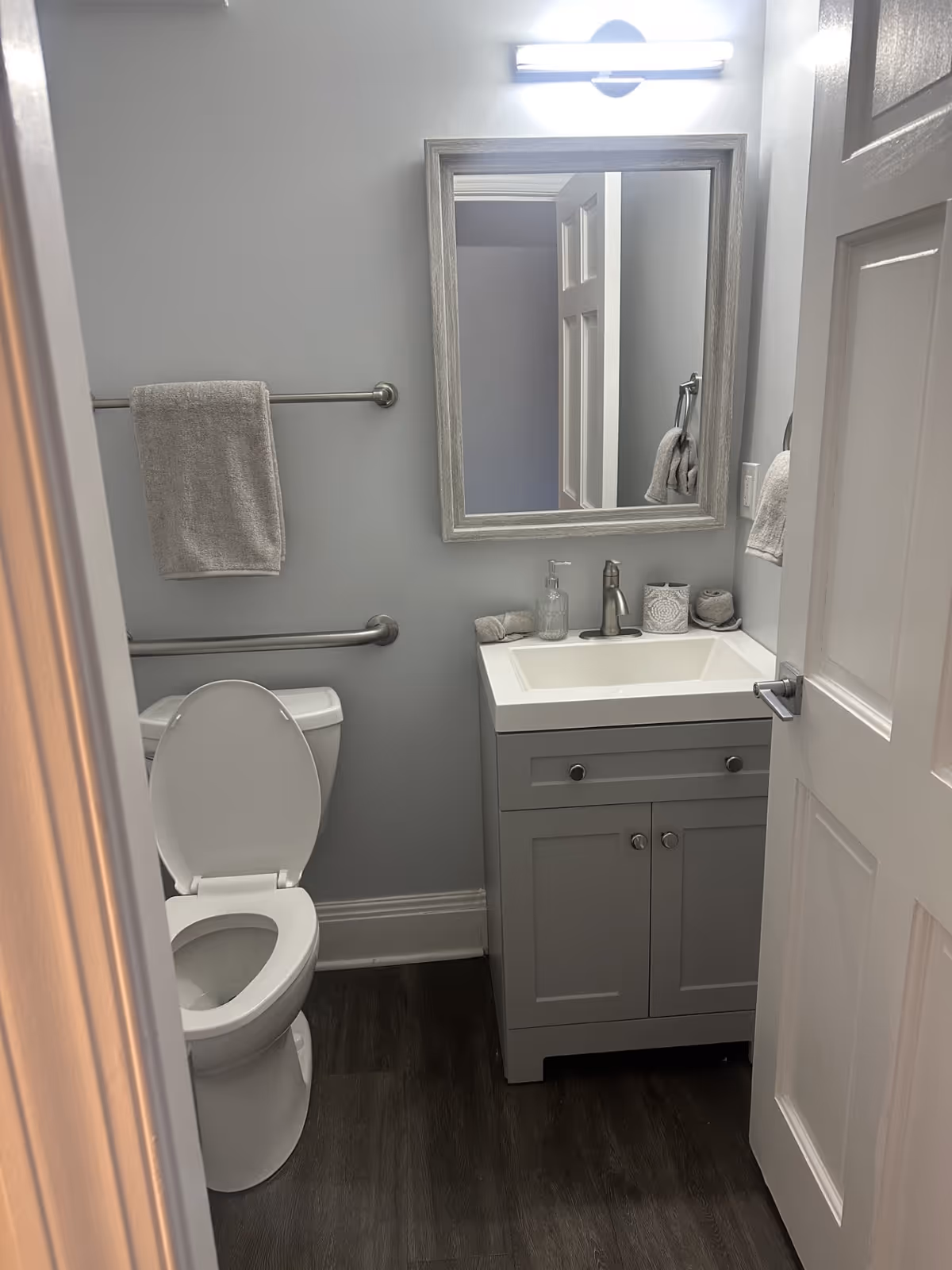 A small, clean bathroom with a white toilet with the lid open, a gray vanity with a white sink, a rectangular mirror above the sink, a towel rack with a gray towel, and a light fixture above the mirror. The floor is dark wood, and the walls are painted light gray.