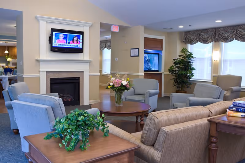 A cozy living room area in a senior living facility with several upholstered chairs and a sofa arranged around a wooden coffee table with a vase of flowers. A flat-screen TV is mounted above a fireplace, and there is a large fish tank built into the wall. The room has large windows with valances, a potted plant, and a side table with a green leafy plant and a stack of books.