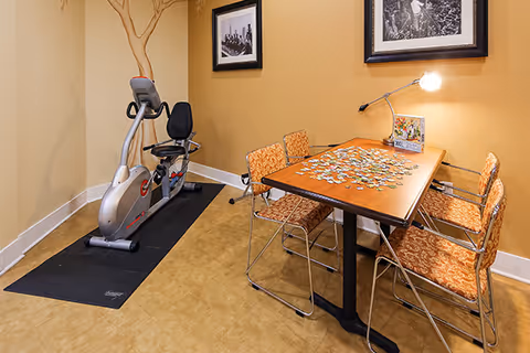 A small room with a stationary exercise bike on a black mat in the corner and a wooden table with four orange patterned chairs around it. The table has a partially completed jigsaw puzzle and a lamp attached to the wall above it. The walls are painted a warm beige color with two black and white framed pictures hanging on the wall.