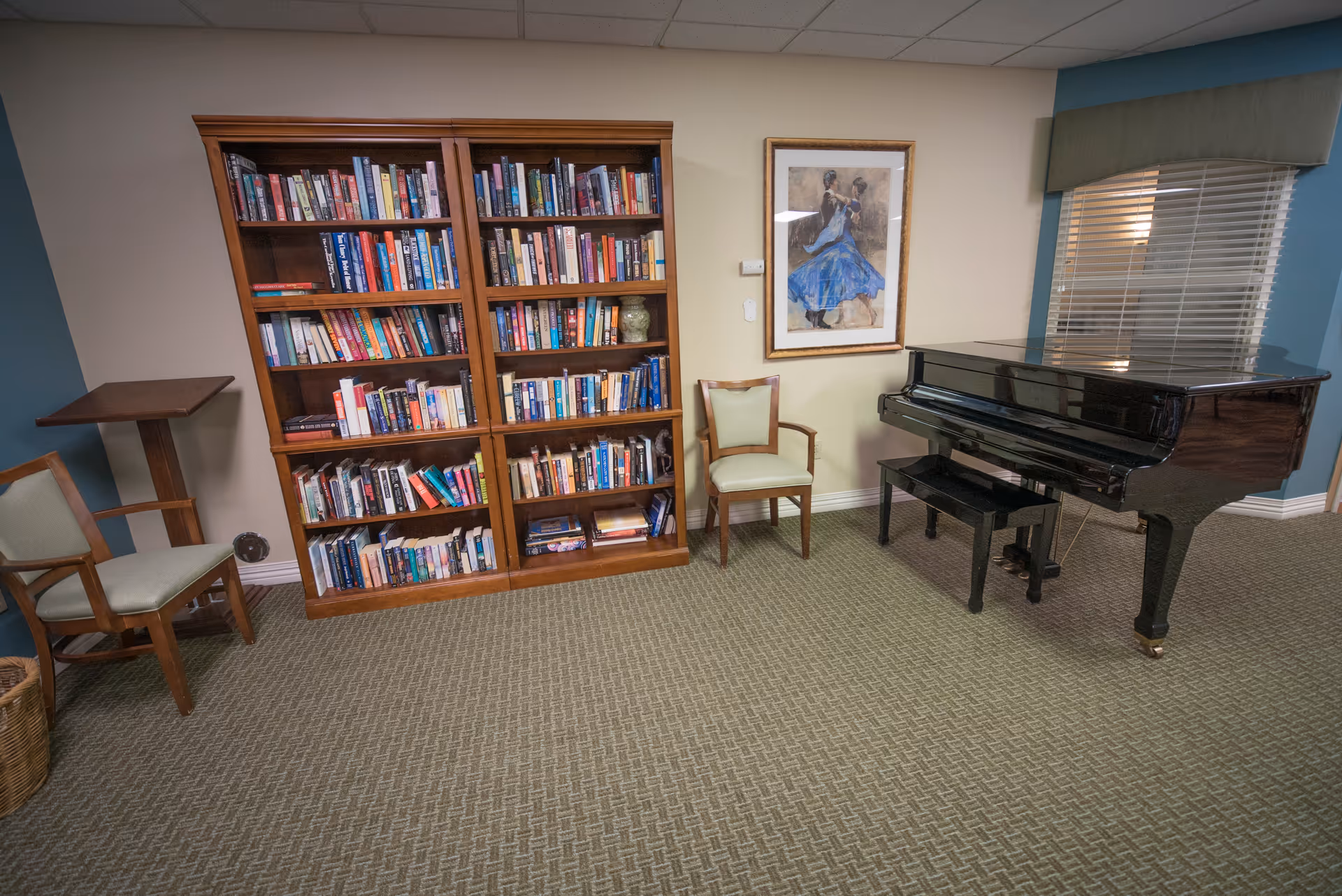 A common room with a large bookcase, chairs, framed artwork and a black grand piano.