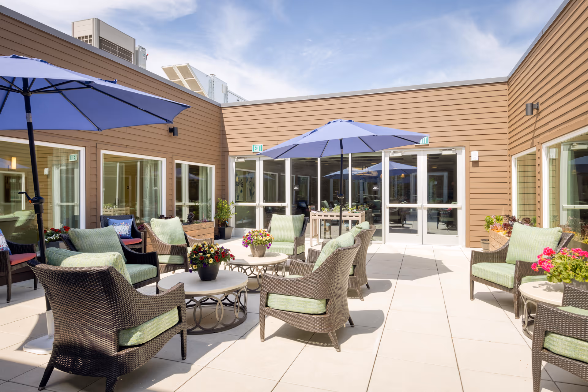 Outdoor patio area with multiple wicker chairs featuring green cushions arranged around small round tables with flower pots. Two large blue umbrellas provide shade. The patio is surrounded by brown siding walls with multiple glass doors and windows, under a partly cloudy sky.