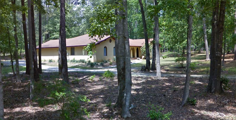A single-story building with a reddish-brown roof and light-colored walls is situated in a wooded area with many tall trees and scattered greenery on the ground.