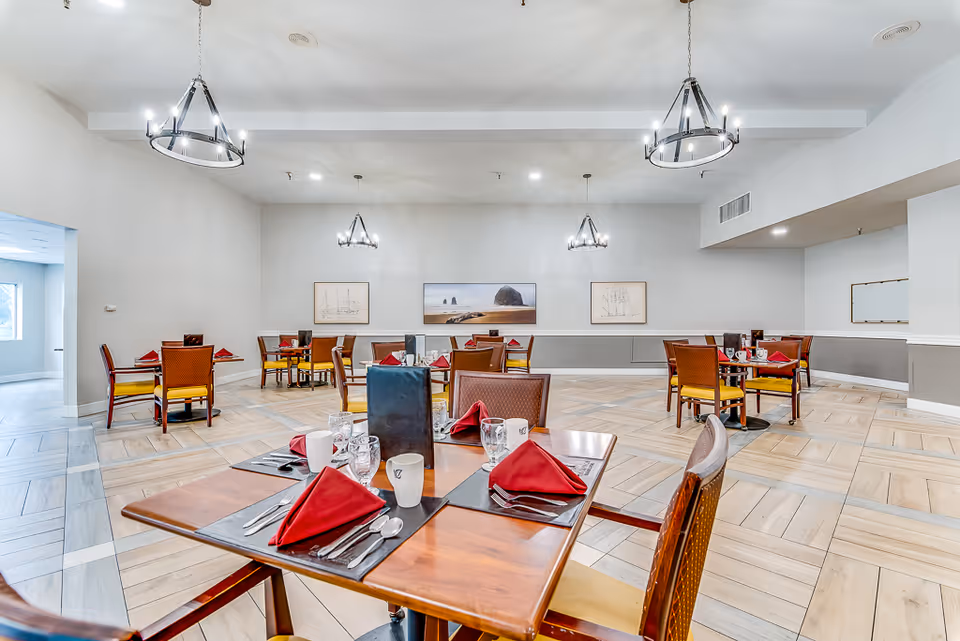 Bright dining room with multiple tables set with red napkins, chandeliers, light wood flooring, and wall artwork.