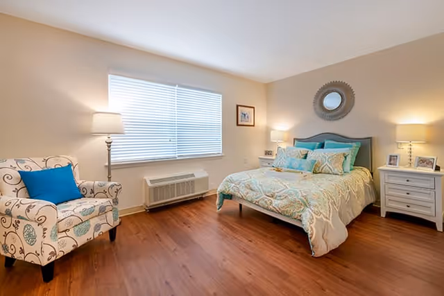 A bright and cozy bedroom with a large window covered by white blinds. The room features a bed with a patterned comforter and multiple pillows, two white nightstands each with a lamp and framed photos, a decorative round mirror above the bed, and a patterned armchair with a blue cushion. The floor is wooden, and the walls are painted a light beige color.
