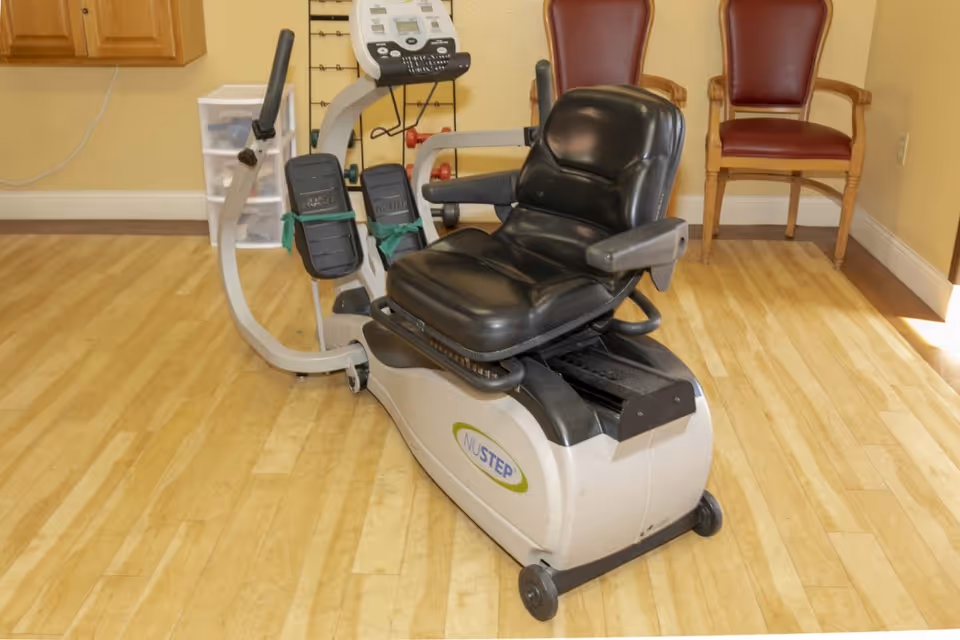A NuStep exercise machine with a black cushioned seat and foot pedals is placed on a wooden floor in a room. In the background, there are two wooden chairs with red cushions and a small plastic drawer unit against a beige wall.