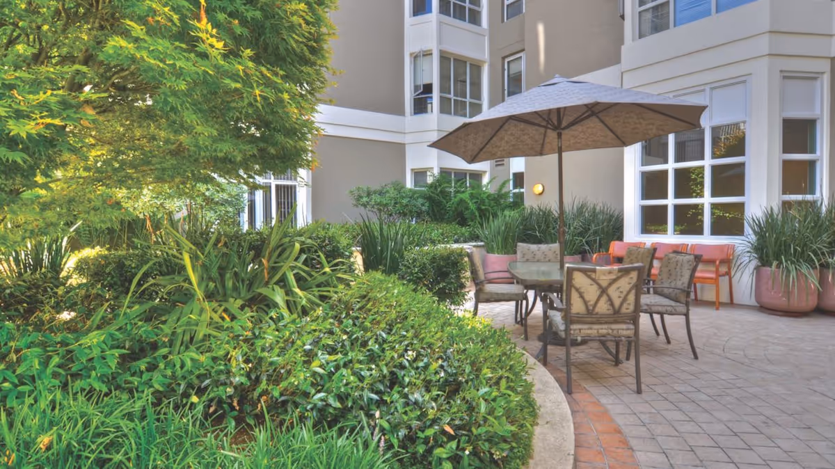 Outdoor patio area at Ivy Park at Cathedral Hill featuring a round table with an umbrella and four cushioned chairs. The patio is surrounded by lush green plants and shrubs, with a beige building and large windows in the background.