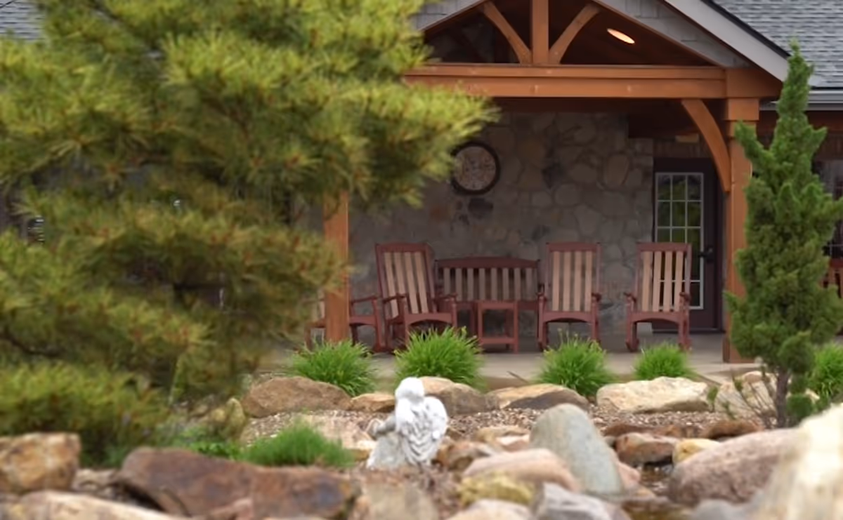 Outdoor seating area with wooden chairs and a bench under a wooden pergola attached to a stone building wall, surrounded by green plants, rocks, and trees in the foreground.