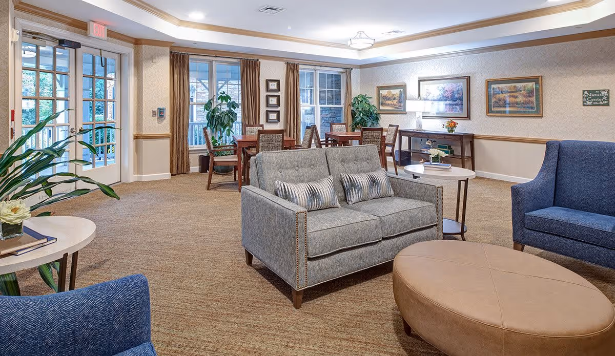 A bright and spacious senior living common area with comfortable seating including a gray loveseat with two striped pillows, a blue armchair, and a large beige oval ottoman. There are several wooden tables and chairs near large windows with brown curtains. The walls are decorated with framed artwork and a small sign that reads 'Nature Center'. Potted plants and a lamp on a console table add to the cozy atmosphere.