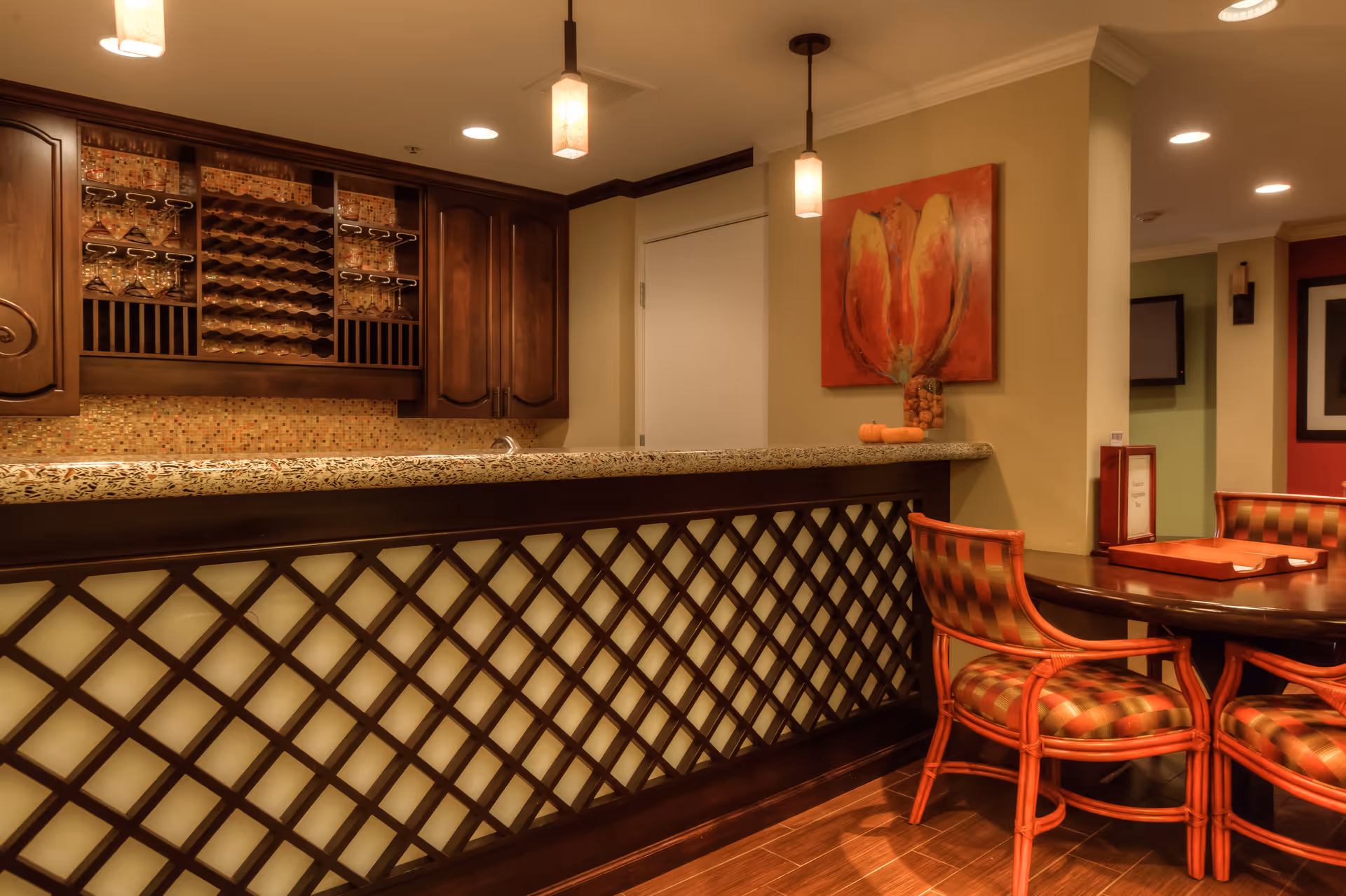 Interior view of a cozy bar area with a granite countertop, wooden cabinets with wine racks and glass holders, pendant lights hanging from the ceiling, and a round table with four cushioned chairs with red and orange plaid upholstery. A colorful painting is mounted on the wall behind the table.