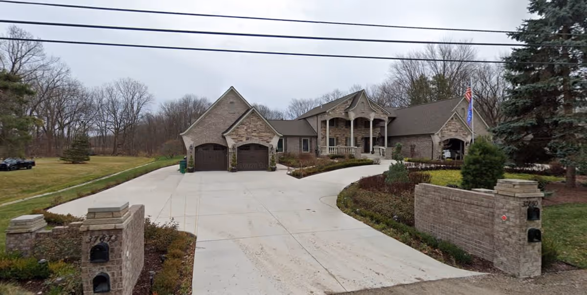 A large, single-story brick house with a wide concrete driveway leading to two garage doors. The house has a covered front porch with columns and is surrounded by landscaped bushes and trees. There are two stone pillars with mailboxes at the entrance of the driveway. The sky is overcast and there are leafless trees in the background.