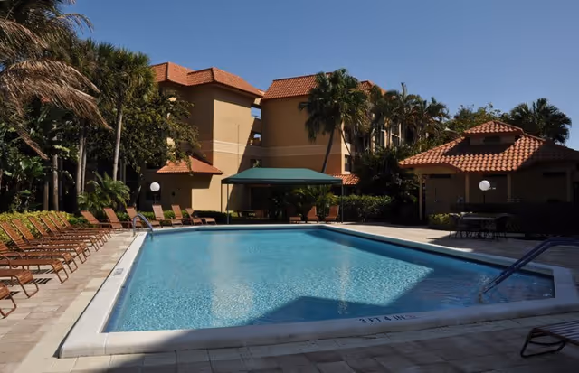 Outdoor swimming pool with lounge chairs, palm trees, and Mediterranean-style buildings with red tile roofs.