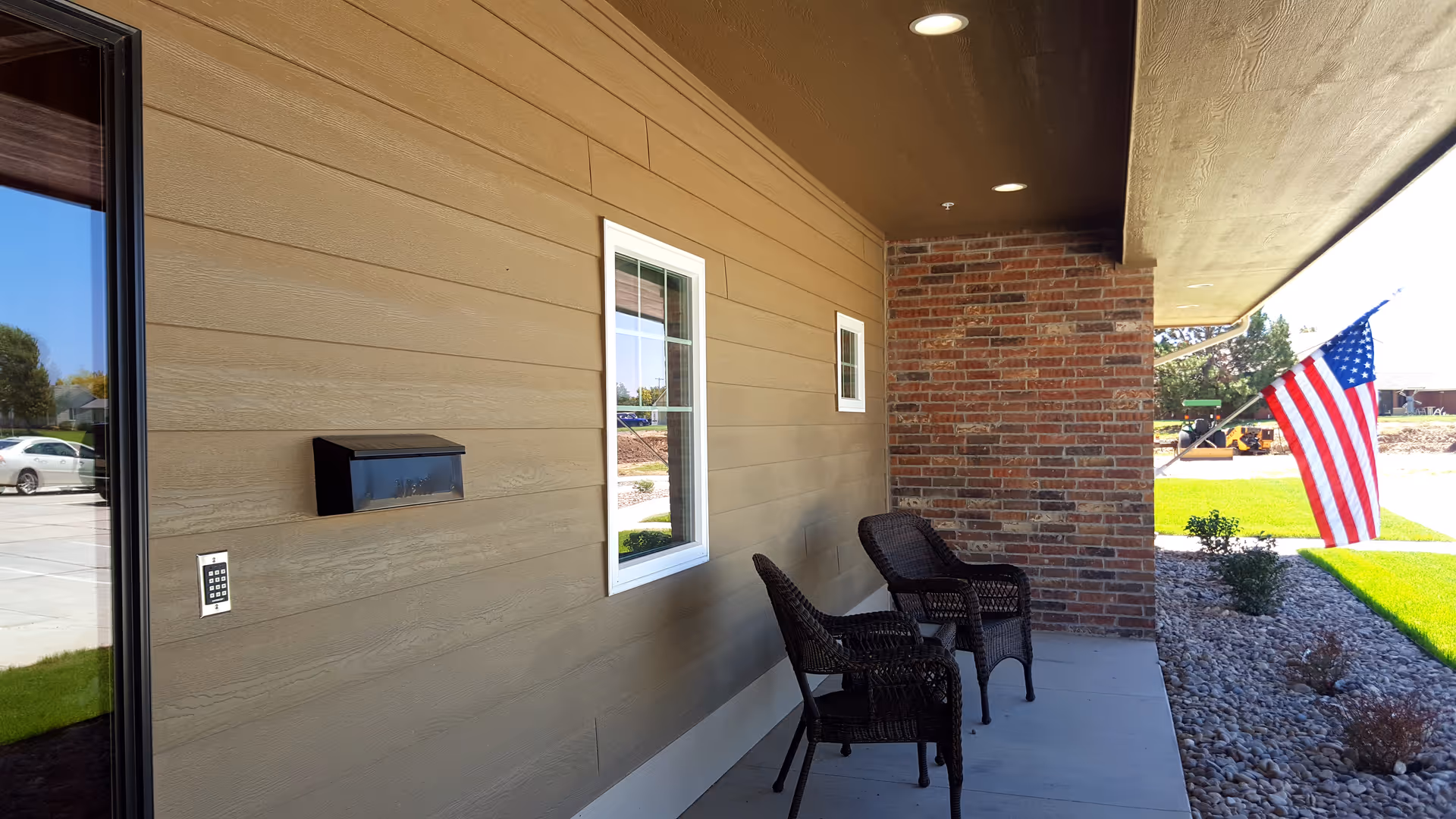 Covered front porch with two wicker chairs, a wall-mounted mailbox and an American flag on the lawn.
