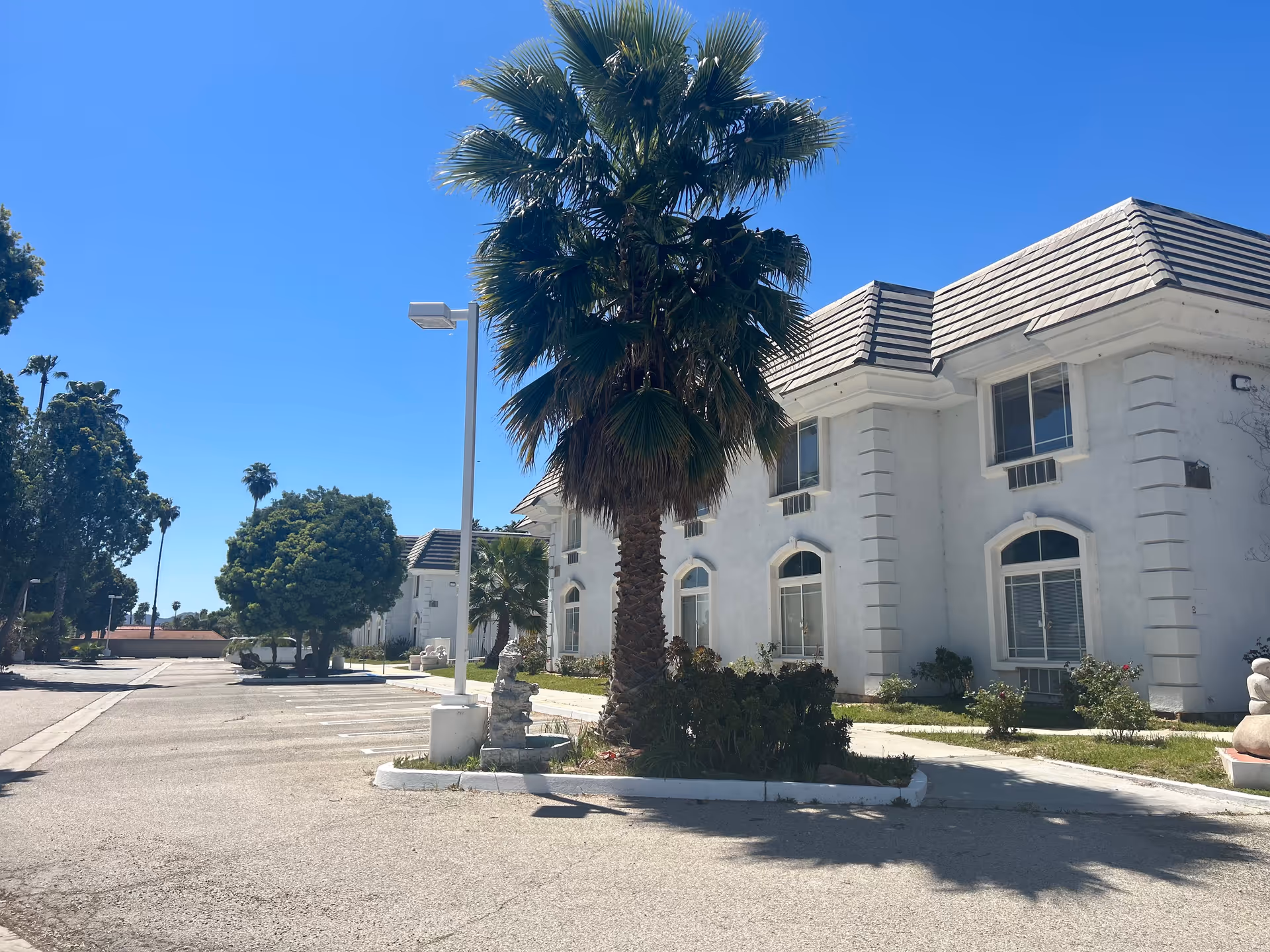 Exterior view of a white two-story building with a tiled roof, surrounded by palm trees and other greenery under a clear blue sky. There is a parking area in front of the building with a street lamp and some sculptures near the sidewalk.