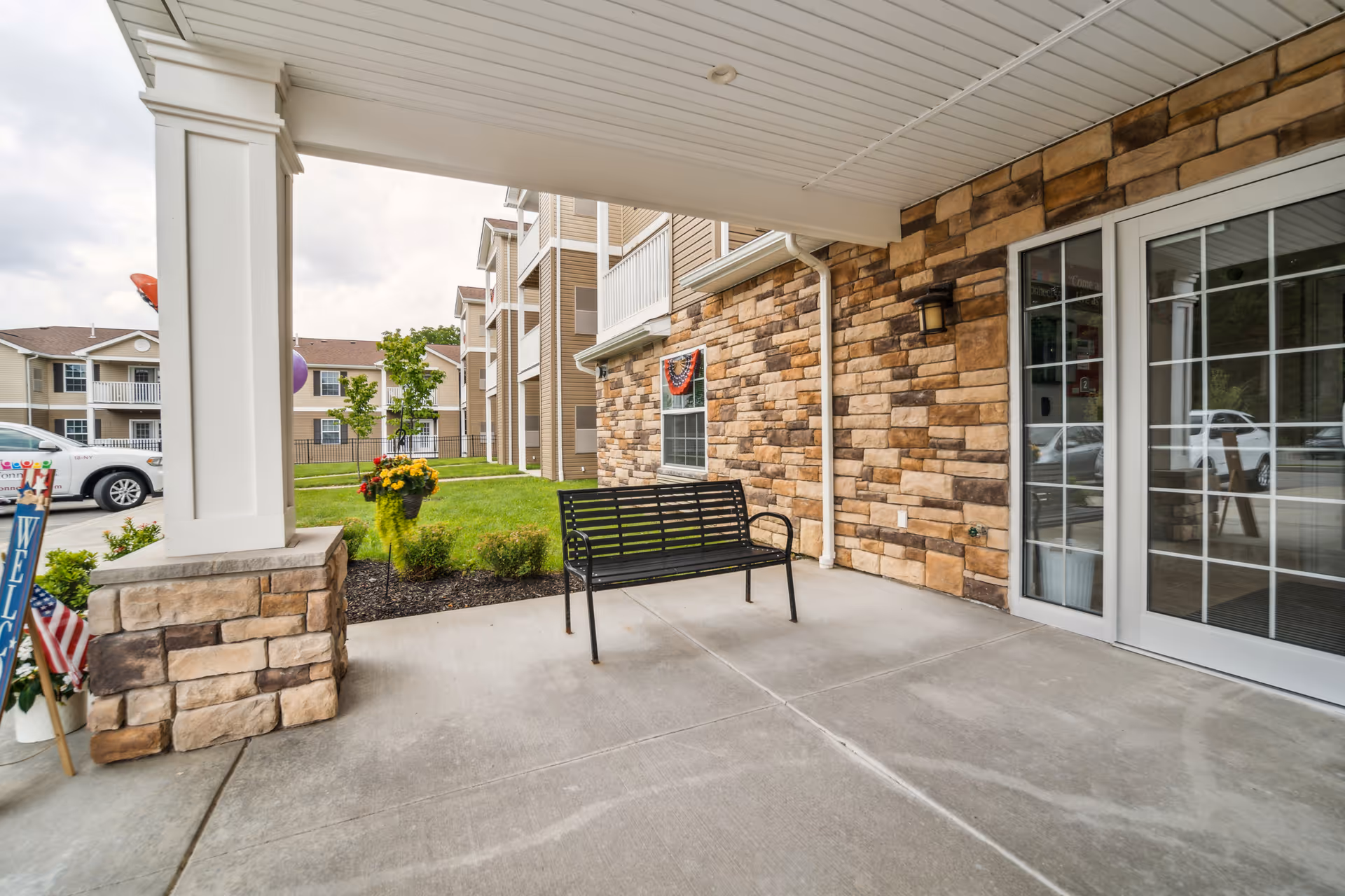 Covered entrance patio of a senior living building with a bench, stone facade, glass doors, and nearby landscaping.