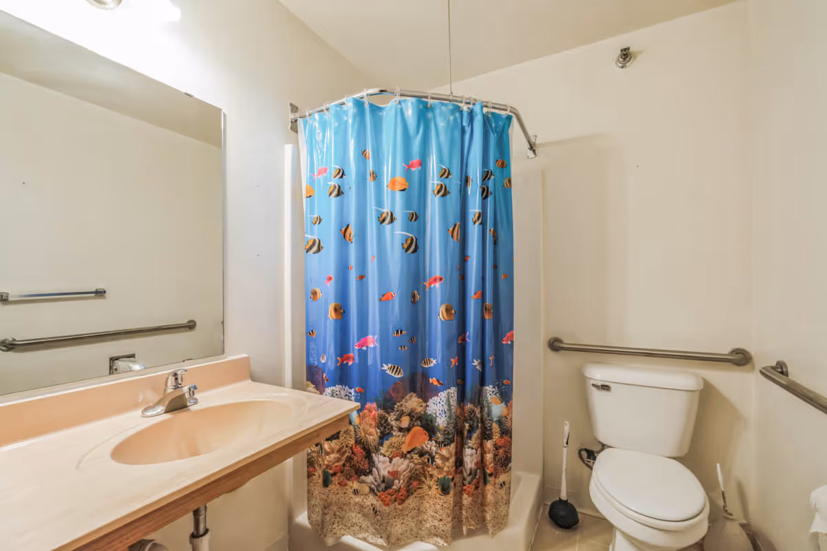 A bathroom with a sink and countertop on the left, a toilet on the right, and a shower with a blue ocean-themed shower curtain featuring colorful fish and coral in the center. The bathroom has grab bars installed near the toilet and along the wall for accessibility.