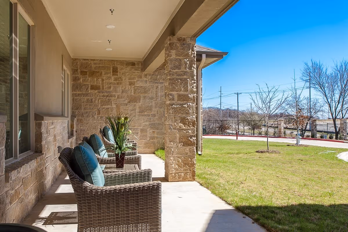 Outdoor covered patio area with wicker chairs featuring blue cushions and a potted plant on a small table. The patio has stone walls and overlooks a grassy lawn with a few small trees and a clear blue sky.