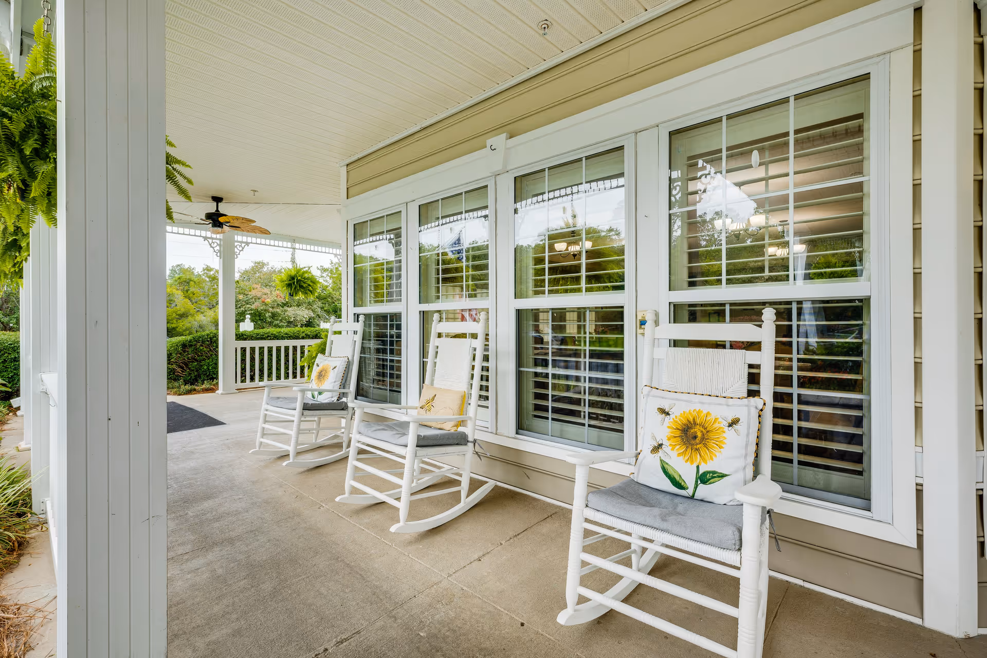 A covered porch area with three white wooden rocking chairs, each with a cushion and a decorative pillow featuring a sunflower design. The porch has large windows with white shutters, hanging green ferns, and a ceiling fan. The surrounding area includes greenery and bushes.