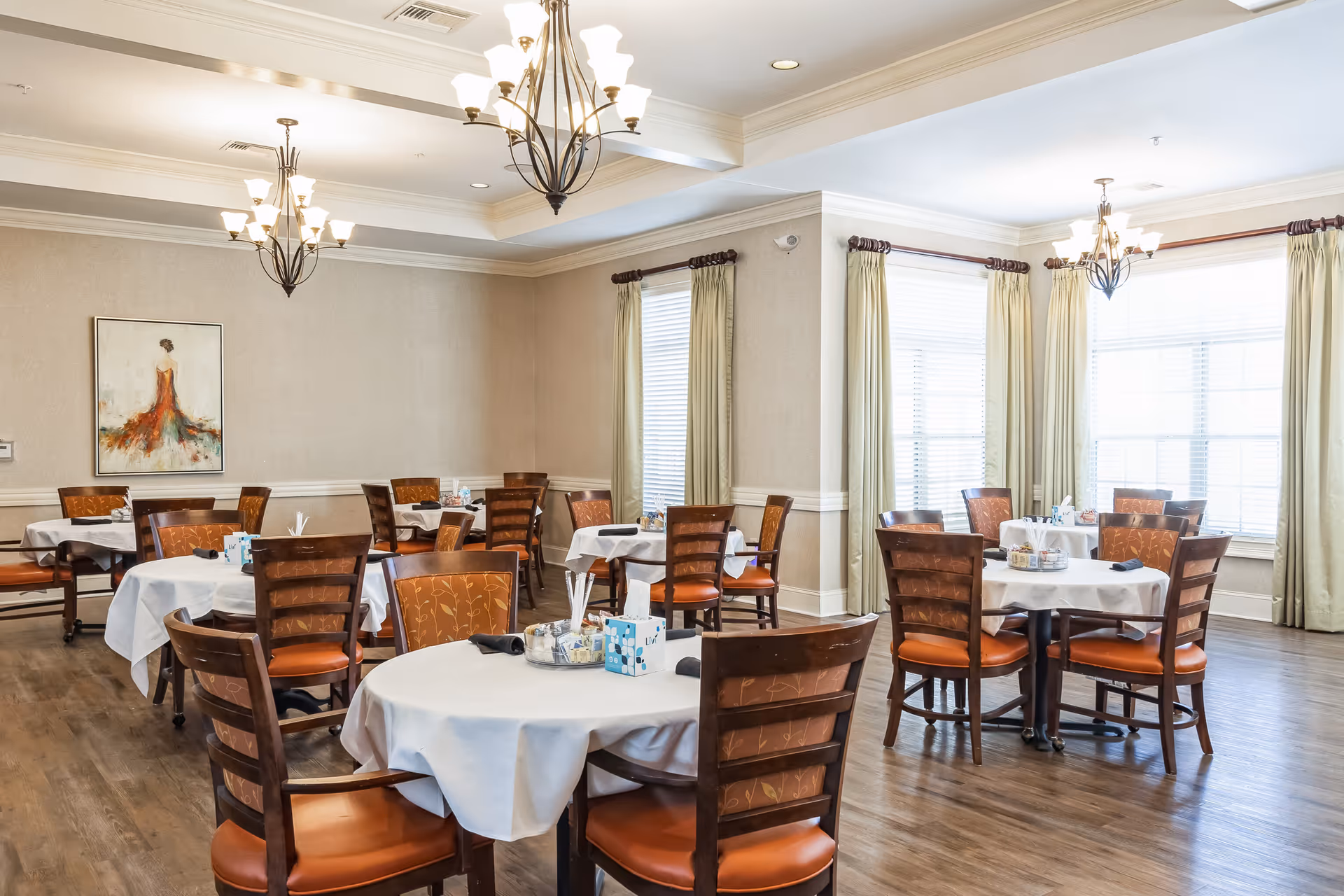 A bright dining room with several round tables covered with white tablecloths, each surrounded by wooden chairs with orange cushions. The room has large windows with cream-colored curtains, wooden flooring, and elegant chandeliers hanging from the ceiling. A painting of a woman in a red dress is mounted on one wall.