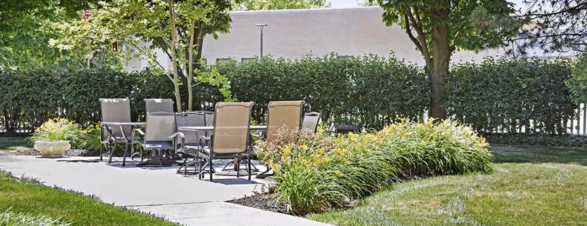 Outdoor patio area with a concrete surface featuring several metal chairs with mesh backs and seats arranged around a table. The patio is surrounded by green grass, bushes, and trees providing shade.