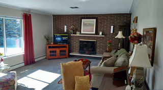 Sunny living room with a large window, brick fireplace, TV on a wooden stand, sofa, chairs, and lamps.
