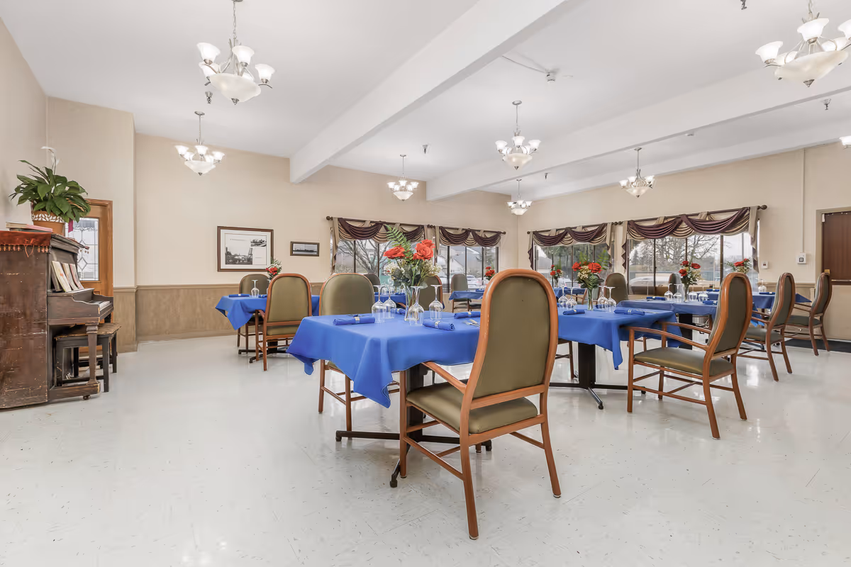 Dining room with multiple tables covered in blue tablecloths, wooden chairs, chandeliers, and a piano against the wall.