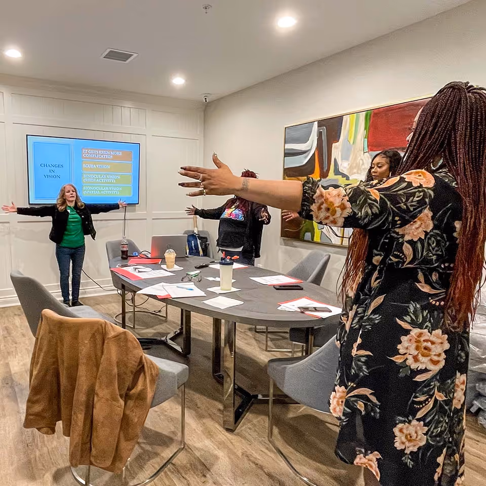 A group of women standing around a conference table in a meeting room, stretching their arms out to the sides. The table has papers, notebooks, and drinks on it. A large screen on the wall displays a presentation slide titled 'CHANGES IN VISION'. The room has modern decor with a colorful abstract painting on the wall.