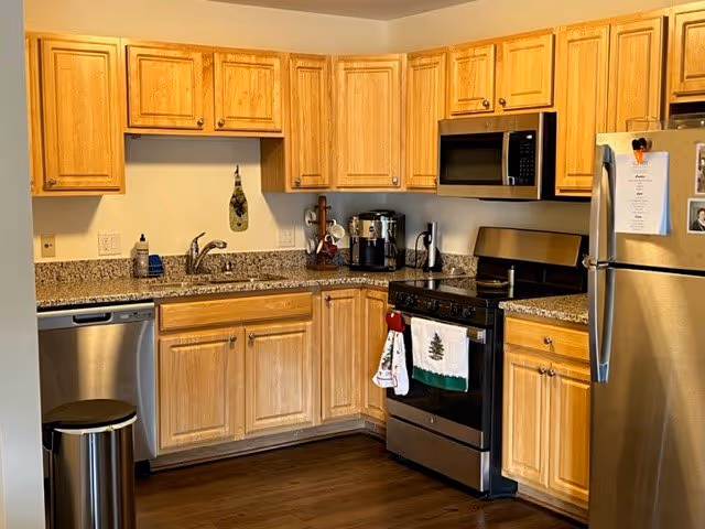 A kitchen with wooden cabinets, granite countertops, and stainless steel appliances including a refrigerator, microwave, dishwasher, and stove. There is a sink with a faucet, a coffee maker, and a trash can. A couple of kitchen towels hang on the oven handle.
