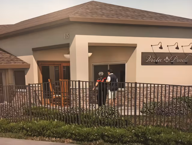 Two people standing outside the entrance of a building with a sign that reads 'Vista Prado'. The building has a beige exterior with stone accents and a brown shingled roof. There is a black metal fence and green shrubbery in front of the building.
