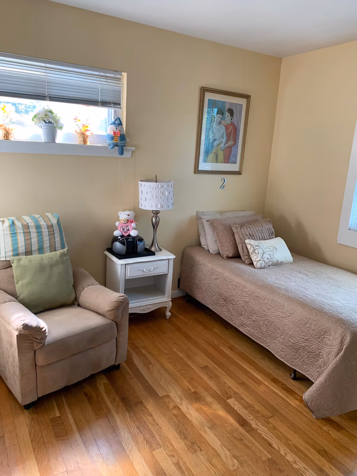 A cozy bedroom with a single bed covered in a beige quilt and several pillows. Next to the bed is a white nightstand with a lamp, a small stereo, and a teddy bear on top. To the left of the nightstand is a beige armchair with a green cushion and a striped pillow. A small window with blinds and flower pots is above the armchair. A framed painting of two people hangs on the wall above the bed, with the number 2 below it. The room has wooden flooring and light beige walls.
