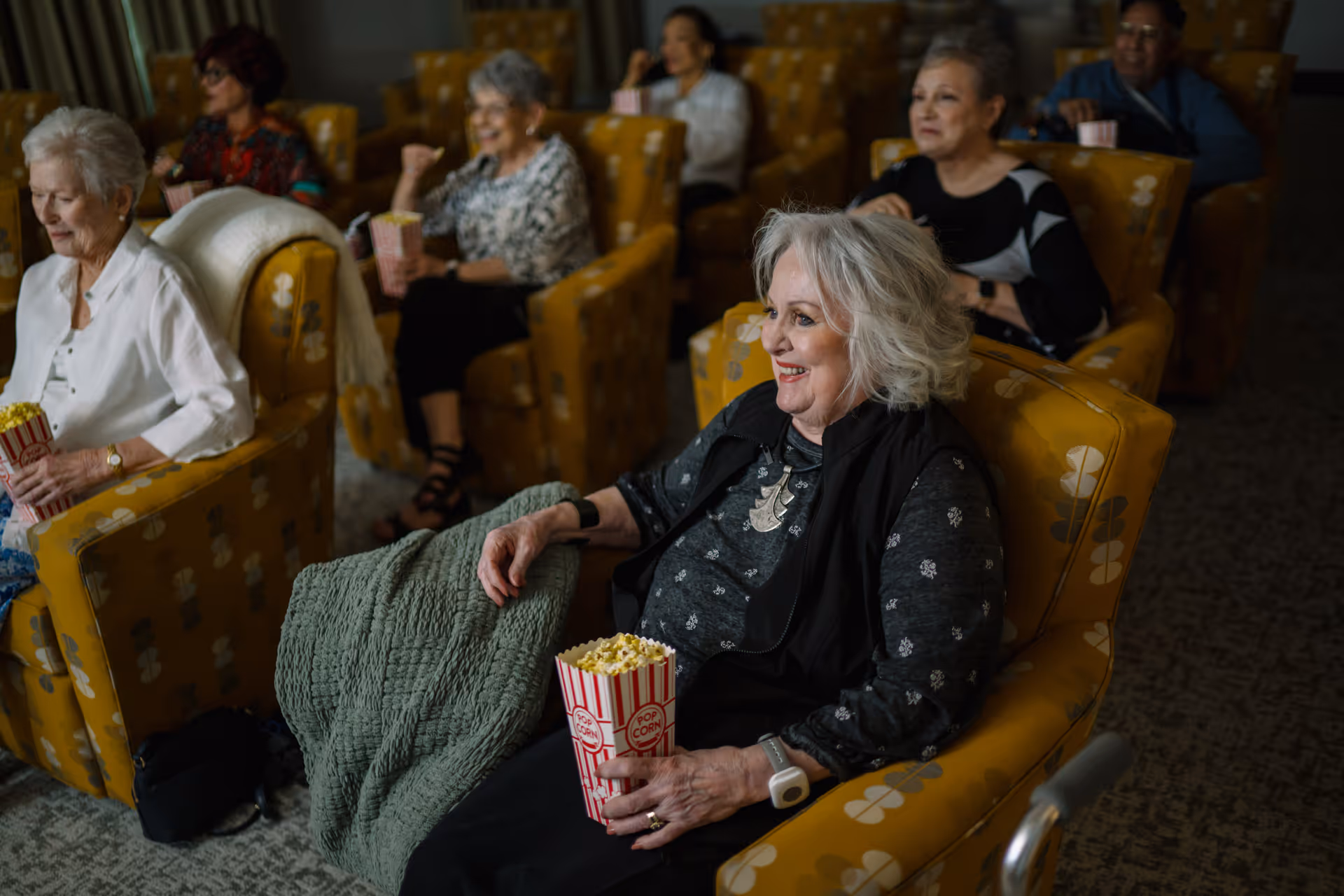 A group of elderly people sitting in comfortable yellow armchairs in a dimly lit room, watching something off-camera while holding popcorn. The atmosphere appears relaxed and social.