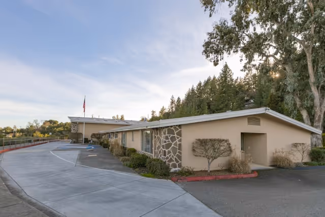 Exterior view of Cloverdale Healthcare Center, a single-story building with beige walls and stone accents, surrounded by trees and greenery under a clear sky.