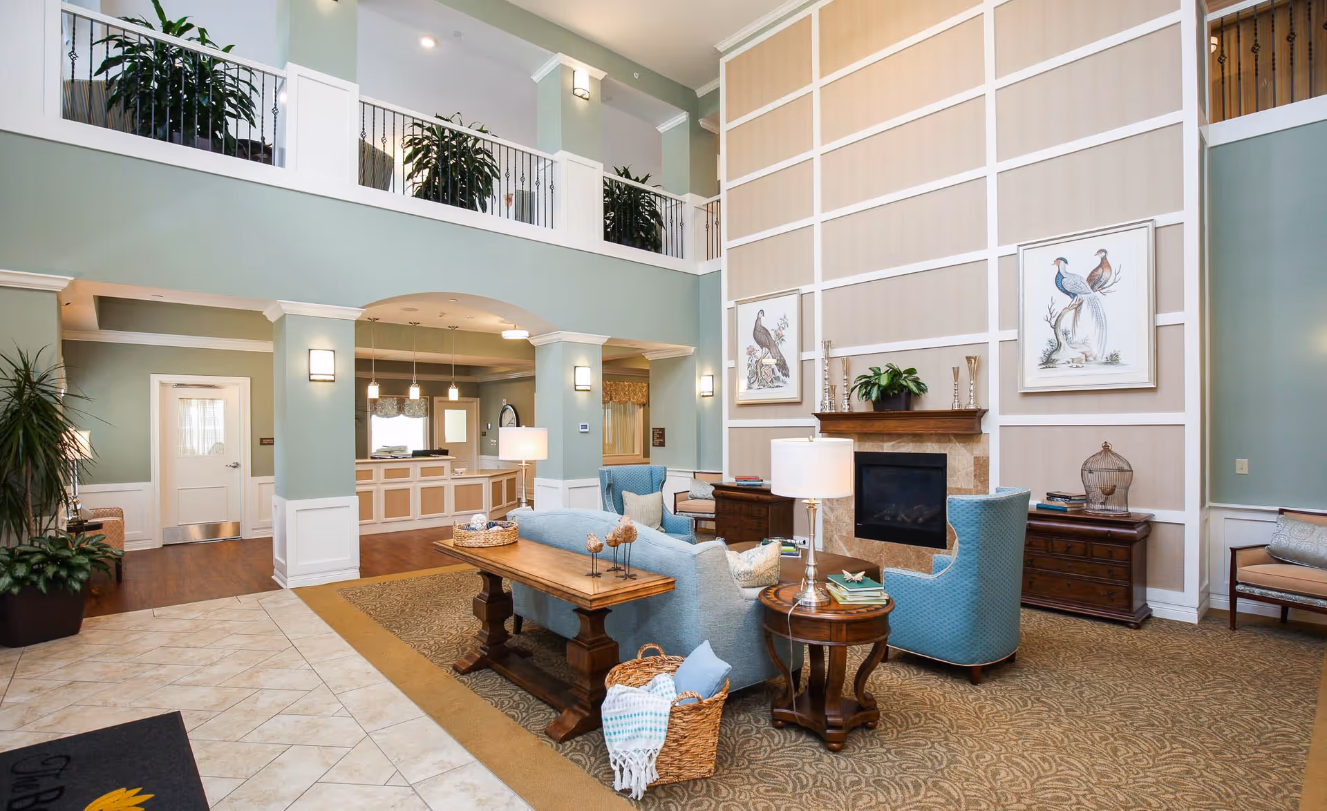 A spacious and well-lit senior living facility common area with high ceilings and a second-floor balcony. The room features comfortable blue armchairs and a sofa arranged around a fireplace with a wooden mantel. There are framed bird illustrations on the wall above the fireplace. The floor is carpeted with a patterned design, and there is a wooden table with decorative items and a basket with a blanket. The walls are painted light green with white trim, and there are several potted plants adding greenery to the space. In the background, a reception desk area is visible under an archway.