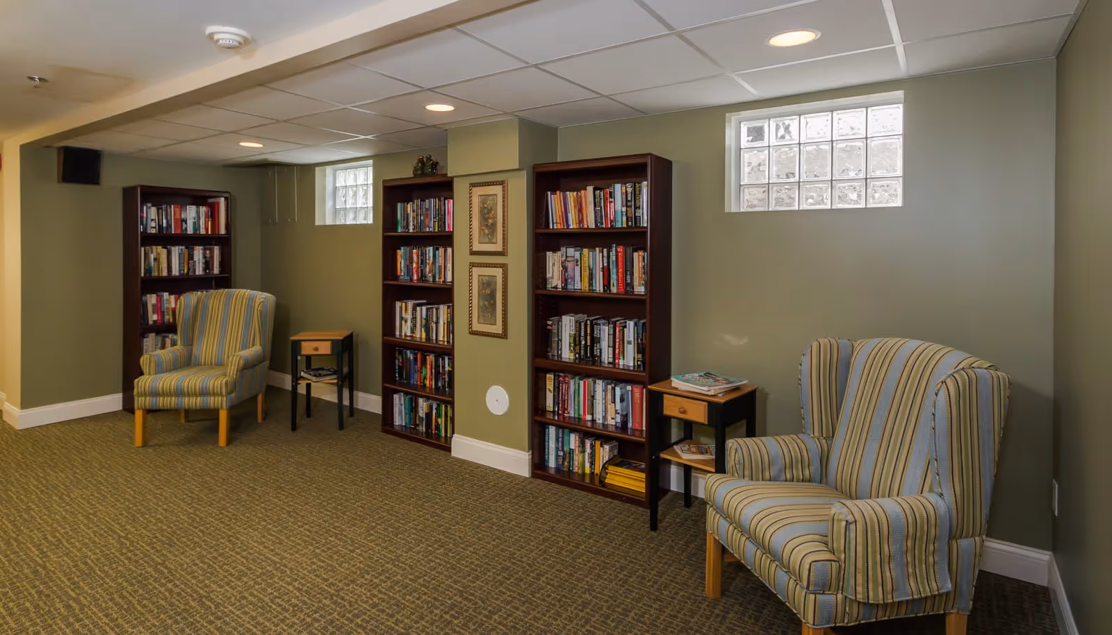 A cozy reading area in a senior living facility with two striped armchairs, three wooden bookshelves filled with books, two small side tables, and two small windows near the ceiling. The walls are painted green and the floor is carpeted.