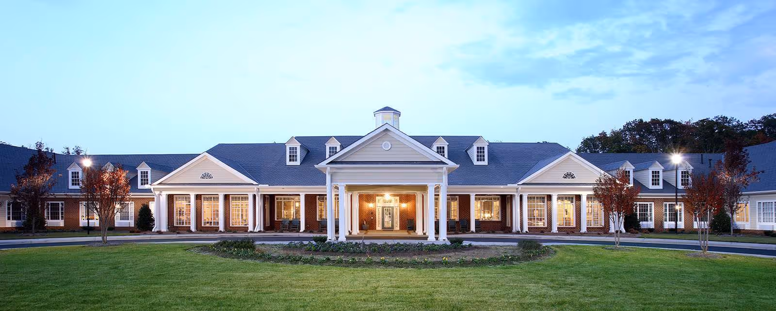 Front exterior view of a large, single-story senior living facility building with white columns, multiple windows, and a well-maintained lawn and driveway in front during dusk.