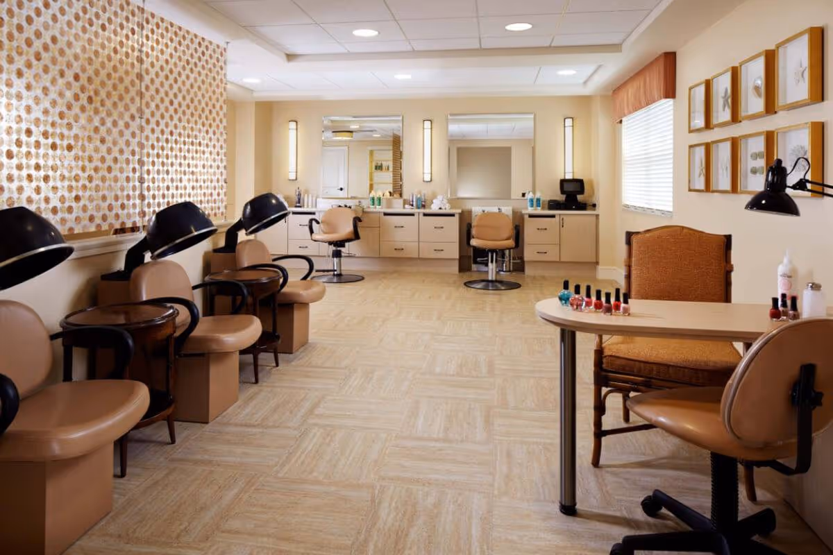 Interior of a salon area in a senior living facility with beige chairs, hair drying stations along the left wall, two styling chairs in front of mirrors on the back wall, and a manicure table with nail polish bottles on the right side. The room has light-colored flooring, soft lighting, and framed artwork on the walls.