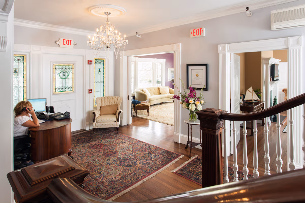 Interior view of a nursing center reception area with a woman sitting at a curved wooden desk on the phone. The space features a chandelier, stained glass windows, a patterned rug, an upholstered armchair, and a small table with a flower arrangement. Adjacent rooms with sofas and decorative items are visible through open doorways.