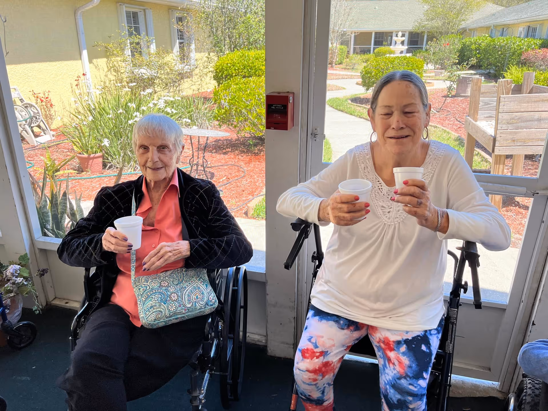 Two elderly women sitting in wheelchairs inside a room with large windows showing a garden outside. Both women are holding white disposable cups and smiling. One woman is wearing a black jacket and coral shirt with a patterned purse on her lap, while the other is wearing a white top and colorful pants.