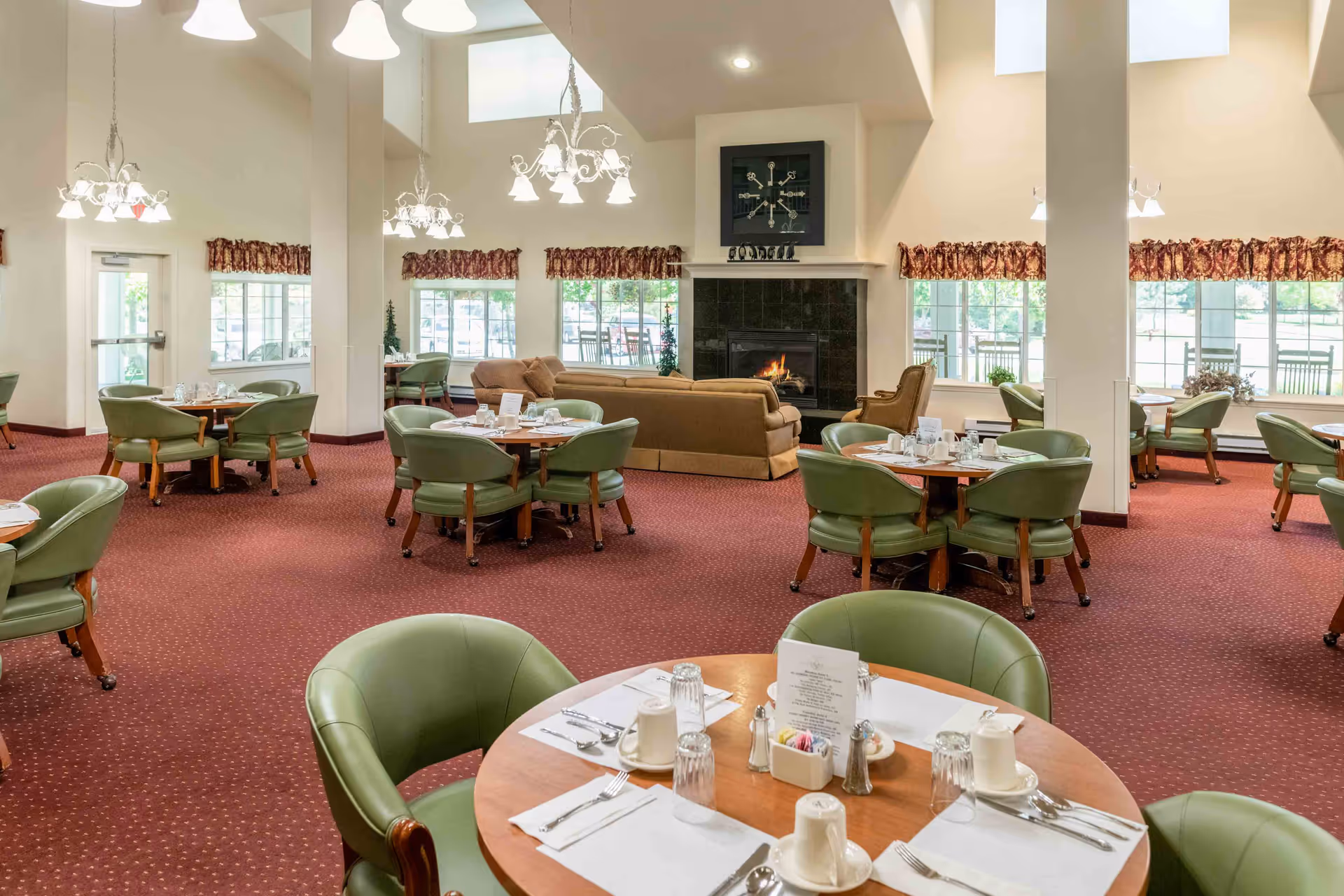 A spacious dining room with multiple round wooden tables set with white placemats, cups, glasses, and silverware. Each table is surrounded by green cushioned chairs on wheels. The room features a red carpet with a subtle pattern, large windows with floral valances, and a cozy fireplace with a fire burning. There are beige sofas and an armchair near the fireplace, and elegant chandeliers hang from the ceiling.