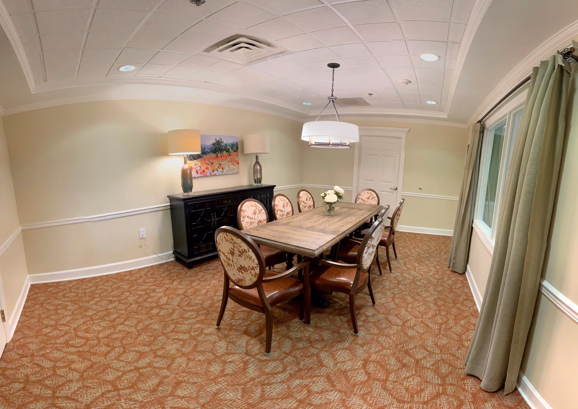 A dining room with a rectangular wooden table surrounded by eight upholstered chairs with floral patterns. The room has beige walls with white trim, a patterned carpet, two table lamps on a black sideboard, a painting on the wall, a hanging ceiling light fixture, and a window with green curtains.