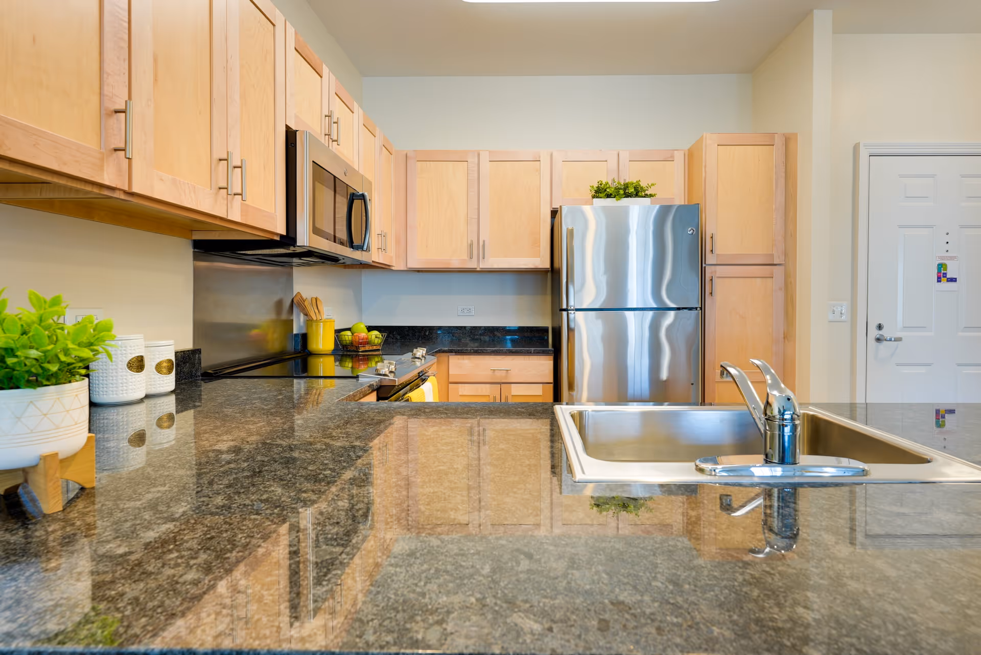 Bright modern kitchen with granite countertops, light wood cabinets, a stainless steel refrigerator and a sink on the island.