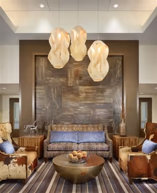 A cozy living room area with a patterned blue sofa and two brown and beige armchairs with blue cushions. A round coffee table with decorative items sits on a striped rug. Three artistic pendant lights hang from the ceiling above the seating area. The back wall features a large textured panel and two side tables with decorative items.