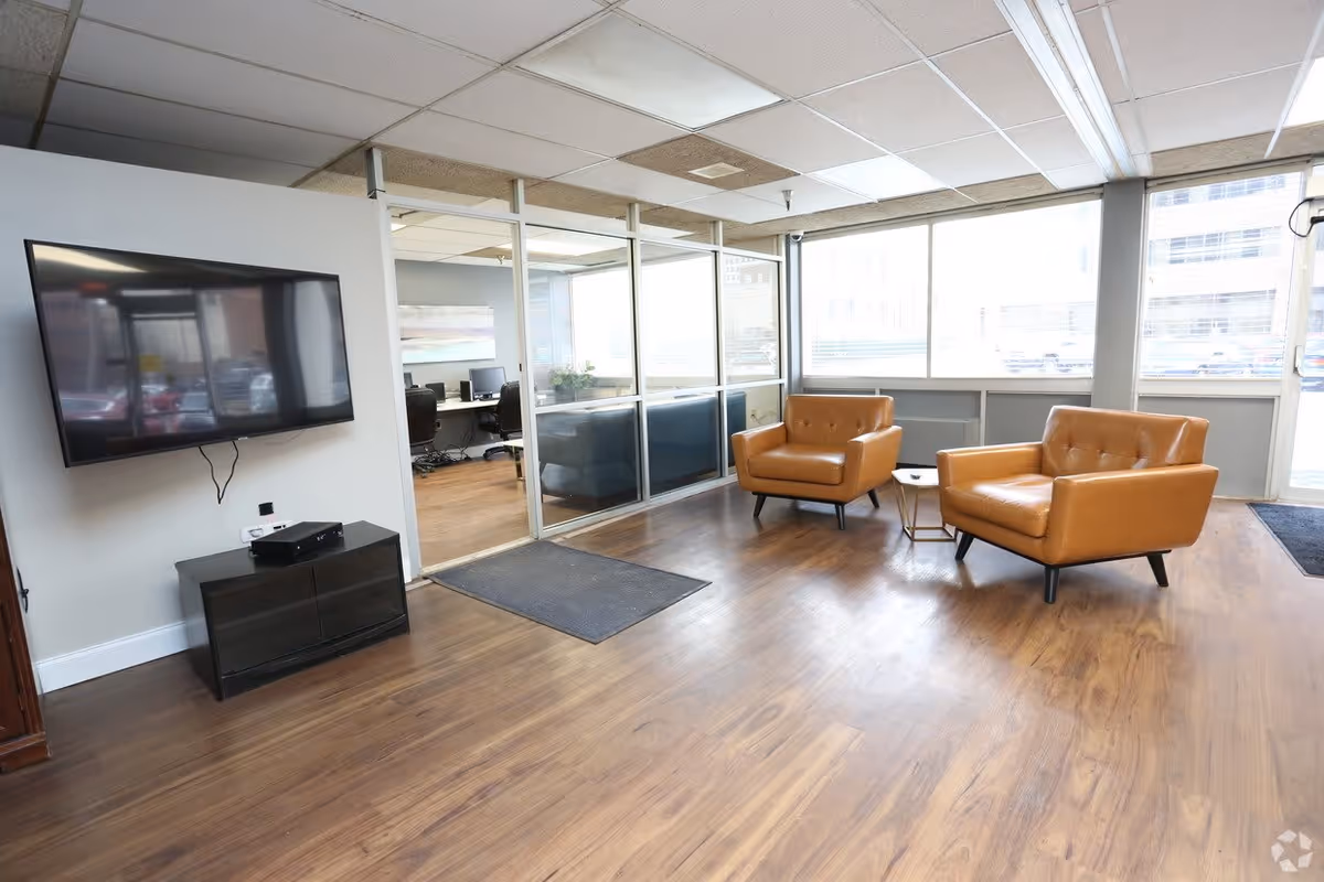 A modern interior lounge area with two tan leather armchairs and a small round side table between them. The room has large windows letting in natural light, a wall-mounted flat-screen TV above a black cabinet, and a glass partition separating the lounge from an office space with desks and chairs.