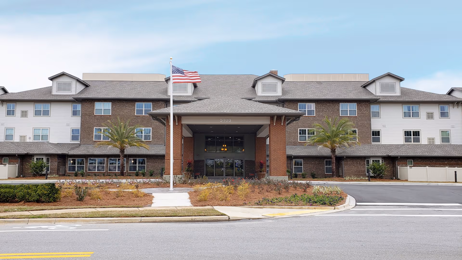 Front exterior view of Red Hills Village Retirement Resort, a three-story building with a combination of brick and light-colored siding. The entrance features a covered porch with columns, and there are two palm trees planted in front. An American flag flies on a flagpole near the entrance, and the surrounding area includes landscaped bushes and a paved driveway.