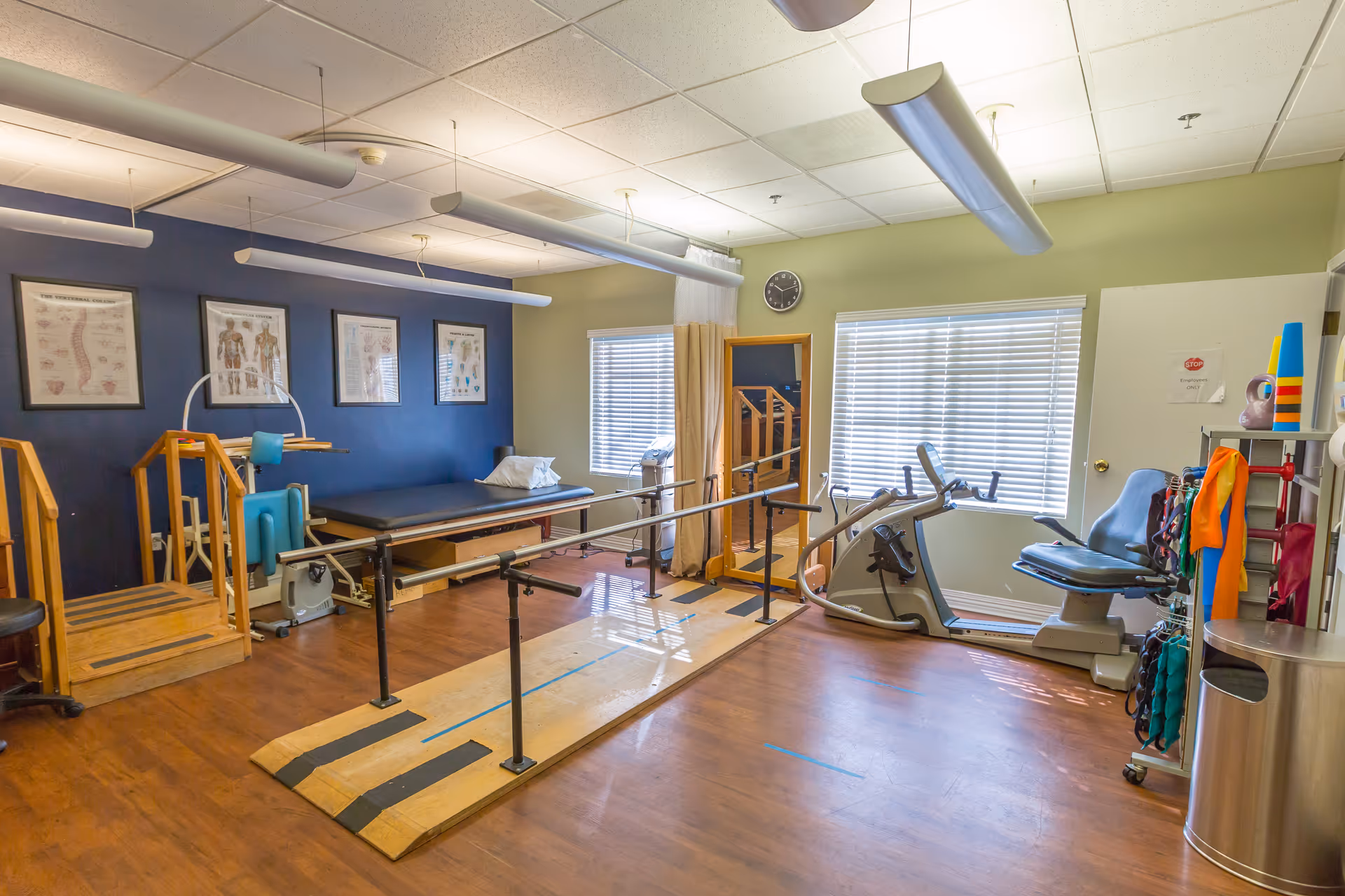 A bright physical therapy room with parallel bars, a treatment table, exercise bike and other rehab equipment under fluorescent lights.
