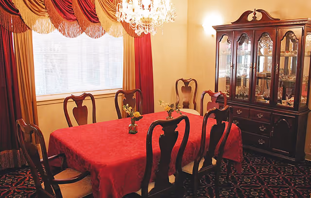 A traditional dining room with a rectangular table covered in a red tablecloth, surrounded by six wooden chairs with cushioned seats. The room features a large window with layered curtains in gold and red, a crystal chandelier hanging above the table, and a dark wooden china cabinet displaying glassware and dishes. The floor is carpeted with a patterned design.