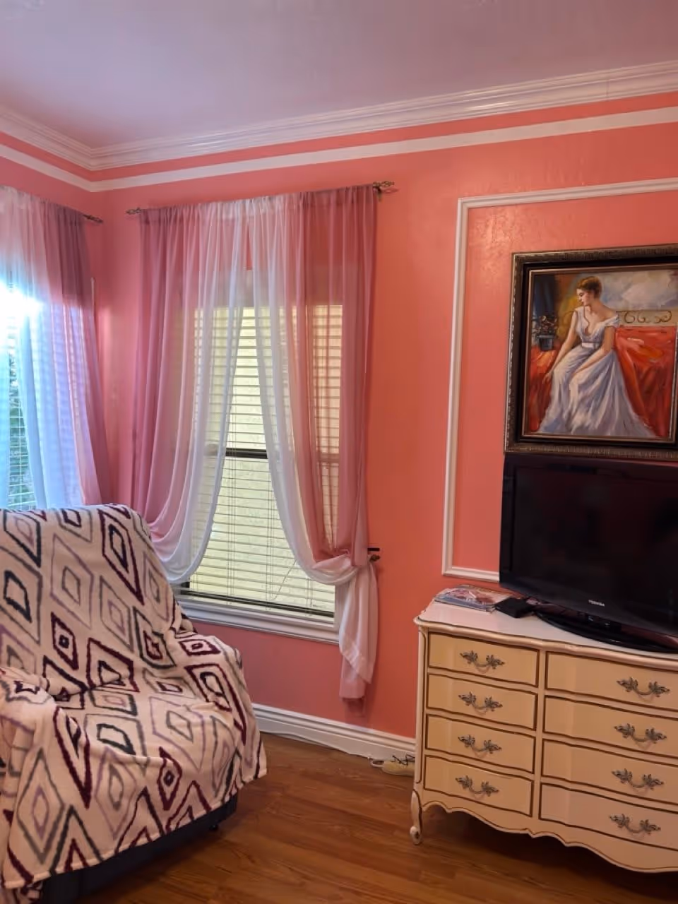 A cozy corner of a room with pink walls and white crown molding. There is a window with white blinds and sheer pink and white curtains. A chair covered with a patterned blanket is positioned near the window. Next to the chair is a cream-colored dresser with ornate handles, on top of which sits a flat-screen TV and some magazines. Above the dresser hangs a framed painting of a woman in a white dress sitting on a red surface.